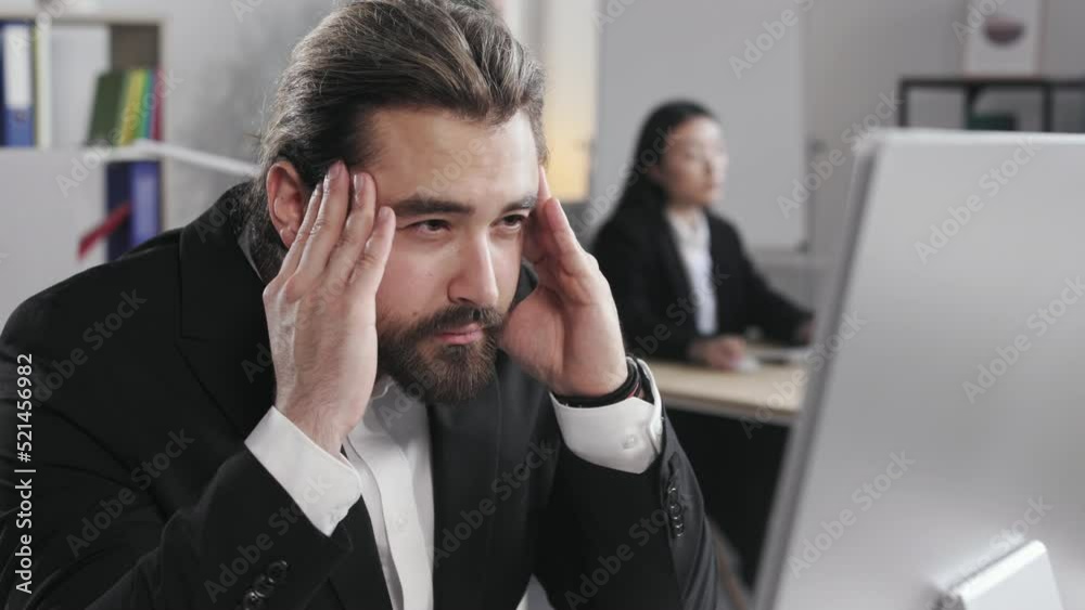 Bearded man with long hair holding hands on temples and looking at computer screen. Young male in formal suit suffering from headache while working at modern office. Bearded man with long hair holding hands on temples and looking at computer screen. Young male in formal suit suffering from headache while working at modern office.