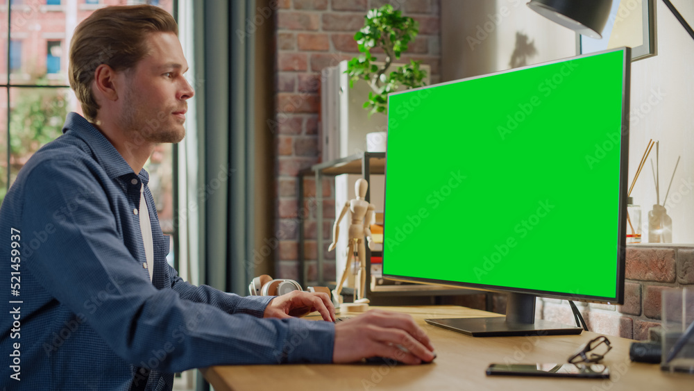 Young Handsome Man Working from Home on Desktop Computer with Green Screen Mock Up Display. Male Checking Corporate Accounts, Messaging Colleagues. Loft Living Room with Big Window.