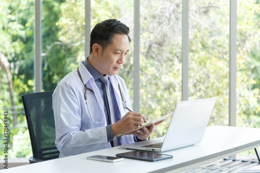 Asian senior doctor sitting in medical office while taking note and using laptop computer. Asian senior doctor sitting in medical office while taking note and using laptop computer.