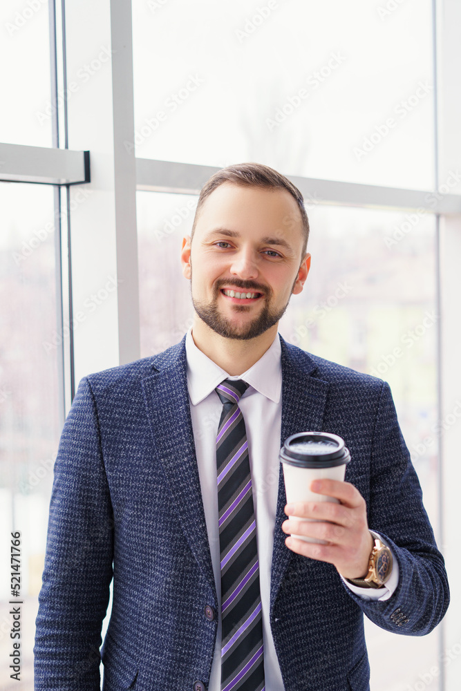 A young man with a neat little beard is dressed in a business suit with a white shirt and tie with a disposable paper cup of tea or coffee by the large window. Positive young businessman at the window