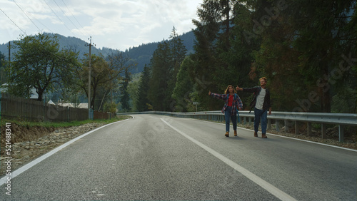 Happy tourists hitchhiking car on highway. Smiling friends wave hands on nature.