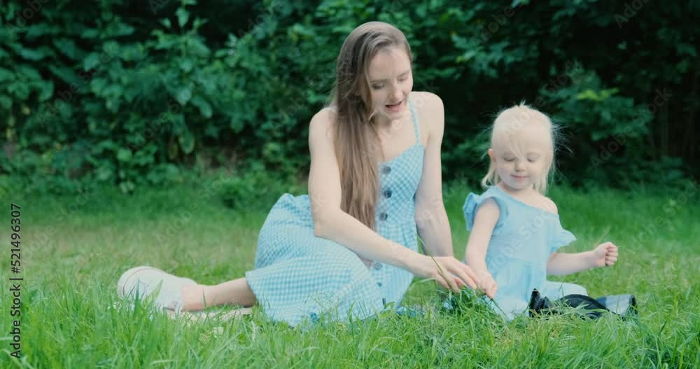 Young woman sits on grass with little blonde girl. Mother with little daughter. Babysitter nanny with child in park Young woman sits on grass with little blonde girl. Mother with little daughter. Babysitter nanny with child in park