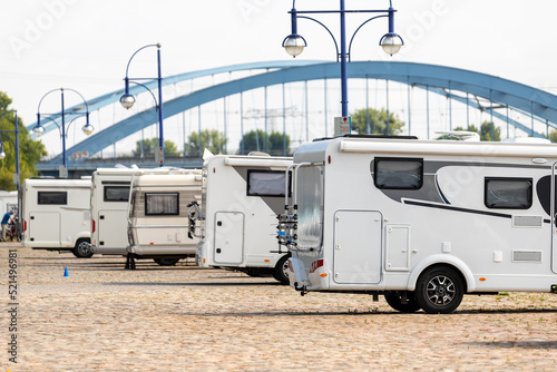 Slika na platnu Many white modern campervan recreational motor home vehicles parked in row at camper park site Magdeburg city against Elbe river bridge