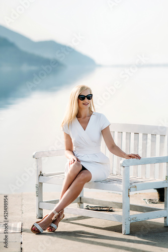 middle aged sexy smiling caucasian woman with sunglasses sits on sea shore in white dress.