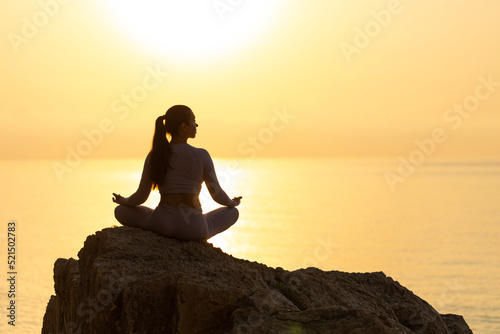 silhouette of young woman practice yoga and meditation near the sea at sunrise, healthy lifestyle concept, lotus position