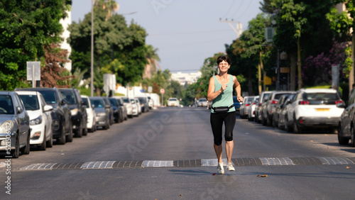 Happy middle aged woman jogging along a street with standing cars on the roadsides in the morning in Israeli city