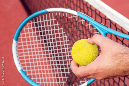 In the men's hand is a tennis racket and a ball against the background a net and tennis court. Close-up image. © Natalia