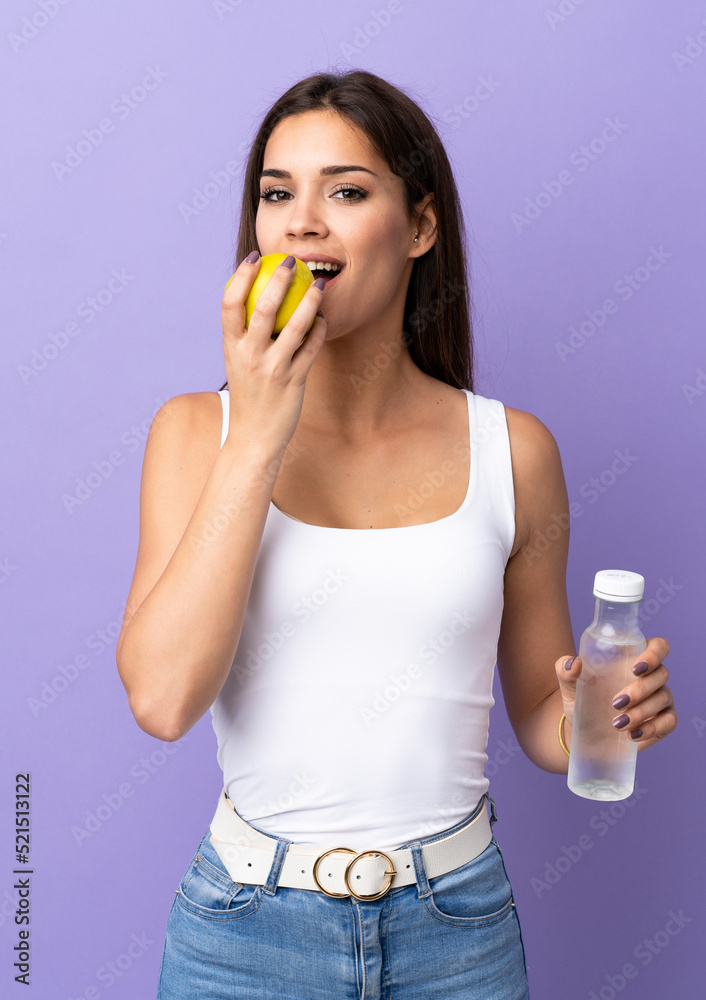 Young caucasian woman isolated on purple background with a bottle of water and eating an apple Young caucasian woman isolated on purple background with a bottle of water and eating an apple