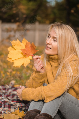 Attractive blonde woman in a yellow sweater with yellow maple leaves in her hands is sitting on a plaid in an autumn park.Autumn concept.Beauty in nature.Slow living.