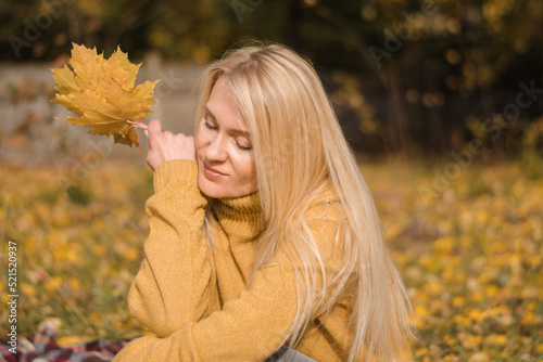 Attractive blonde woman in a yellow sweater with yellow maple leaves in her hands is sitting on a plaid in an autumn park.Autumn concept.Beauty in nature.Slow living.
