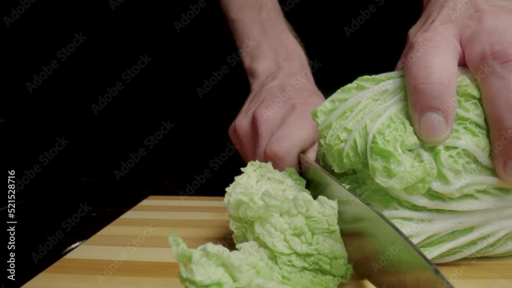 Male hands cutting Chinese cabbage or green lettuce leaves with sharp knife. Chef slicing leafy greenery on wooden cutting board. Cooking in the kitchen or in a restaurant. Black background. Close up. Male hands cutting Chinese cabbage or green lettuce leaves with sharp knife. Chef slicing leafy greenery on wooden cutting board. Cooking in the kitchen or in a restaurant. Black background. Close up.