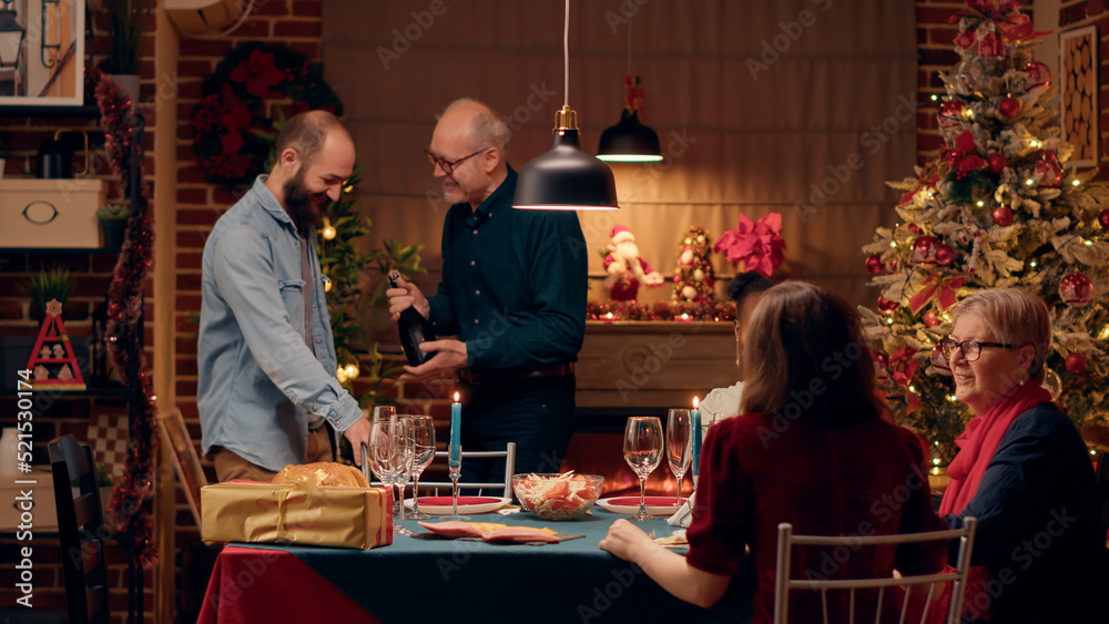 Laughing heartily senior man talking with young adult about champagne while enjoying Christmas dinner. Festive diverse people celebrating winter holiday with traditional authentic home cooked food.