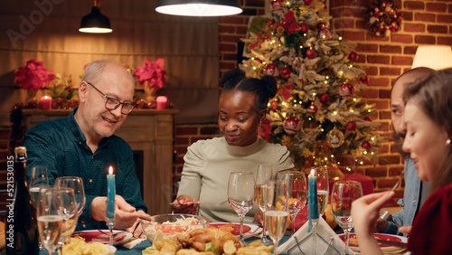 Diverse festive people sitting at Christmas dinner table while enjoying seasonal home cooked food. Happy family members gathered at home in living room while celebrating traditional winter holiday.