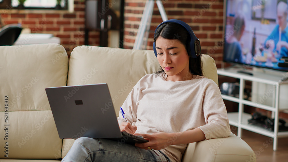 Attentive worker doing remote work on laptop while writing down important job informations. Focused woman taking notes while working remotely and listening to music at home in living room. Tripod shot Attentive worker doing remote work on laptop while writing down important job informations. Focused woman taking notes while working remotely and listening to music at home in living room. Tripod shot