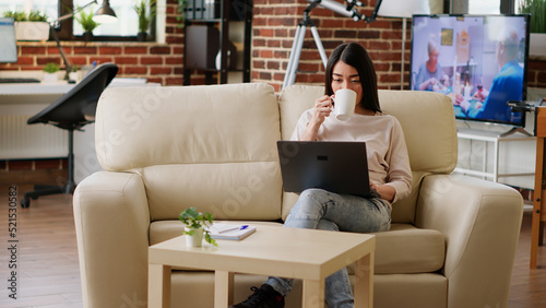 Smiling young adult freelancer enjoying remote work while sitting on couch inside modern flat. Happy asian woman sipping beverage while working remotely on portable computer at home.