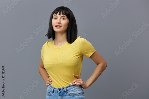 a happy, beautiful, attractive woman stands in a yellow T-shirt and light jeans on a gray background, smiling pleasantly and holding her hands on her belt. Studio Photography