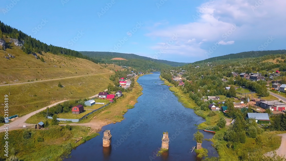 Bird's-eye view. Clip. View of the river near the forest and behind the old mountains, bare sky and small houses around the river