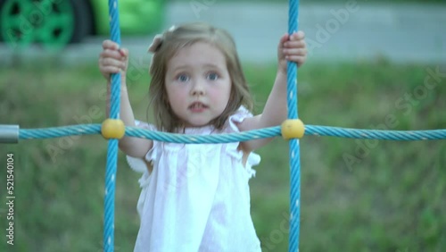 Three-year-old girl in a white dress on the playground. Cute little girl climbing on rope mesh at playground. Child climbing a rope ladder