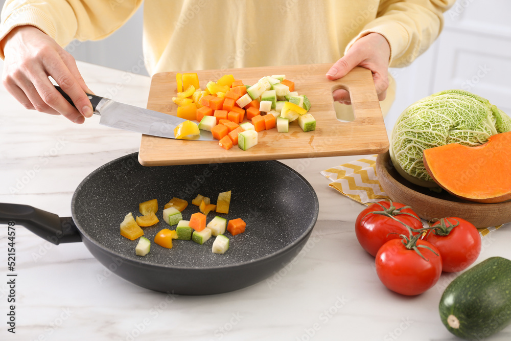 Woman pouring mix of cut vegetables into frying pan at table in kitchen, closeup