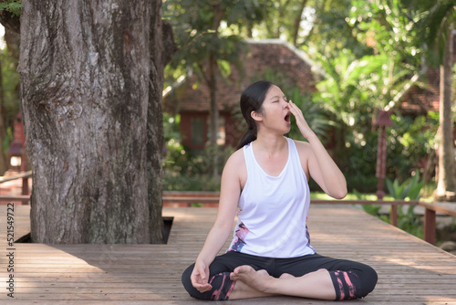 Outdoor portrait of young Asian woman sitting under big tree, being bored and sleepy, yawning while practicing yoga. Laziness concept.