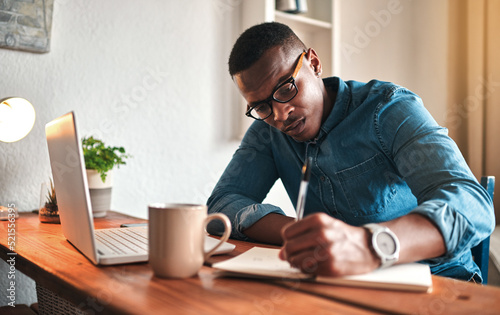 African american entrepreneur writing in a notebook and planning a project strategy. Young man remote working from home with a laptop at his startup business while scheduling appointments in a book