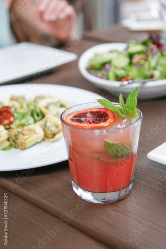 A view of a red colored mint and blood orange garnished cocktail, among several assorted restaurant entrees.