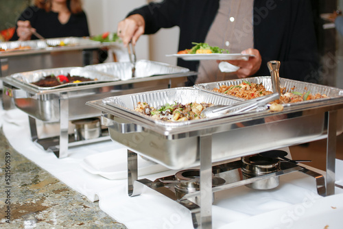A view of several chafer dishes filled with savory entrees, seen at a local catered event. Guests are seen serving themselves. photo