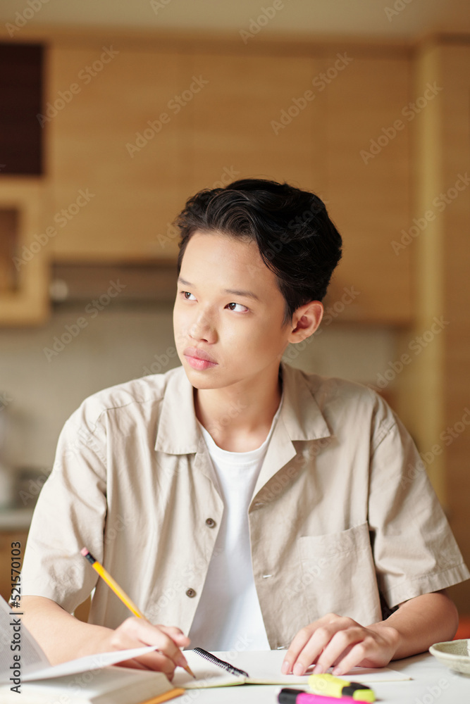 Portrait of pensive teenage boy looking outside through kitchen window when doing homework at home