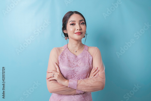Portrait of a confident smiling girl standing with arms folded and looking at camera isolated over blue background, wearing modern kebaya dress