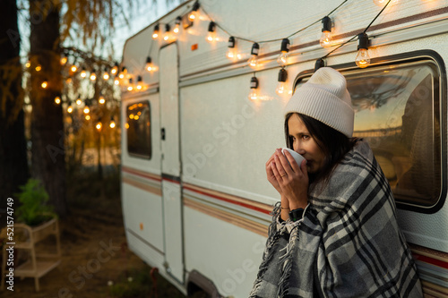 Caucasian woman in a knitted hat wrapped in a plaid and drinks a warming drink outdoors. Travel in a motor home in the fall.