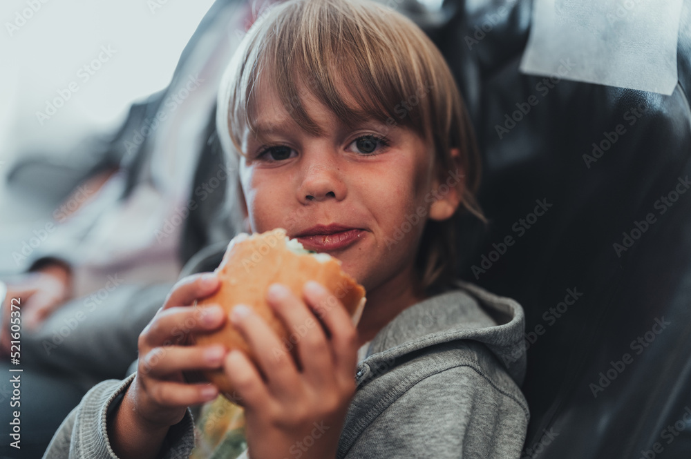 little candid kid boy five years old eats burger or sandwich food sitting in airplane seat on flight traveling from airport. children take a bite. child in air plane eating lunch or dinner meal little candid kid boy five years old eats burger or sandwich food sitting in airplane seat on flight traveling from airport. children take a bite. child in air plane eating lunch or dinner meal