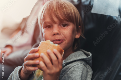 little candid kid boy five years old eats burger or sandwich food sitting in airplane seat on flight traveling from airport. children take a bite. child in air plane eating lunch or dinner meal. flare