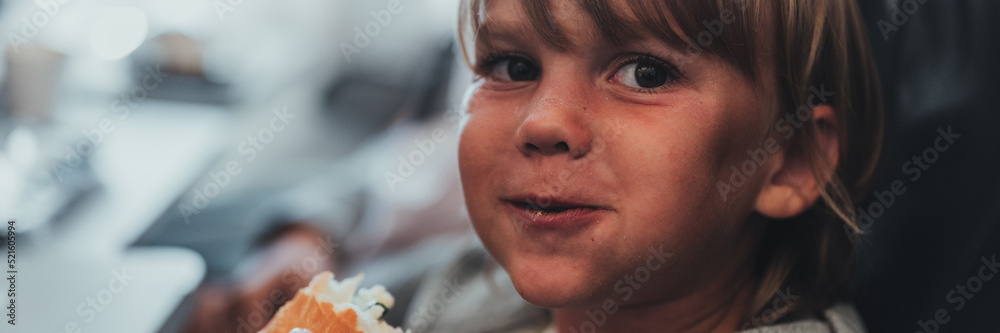 Obraz premium little candid kid boy five year old eats burger or sandwich food sitting in airplane seat on flight traveling from airport. children take a bite. child in air plane eating lunch or dinner meal. banner