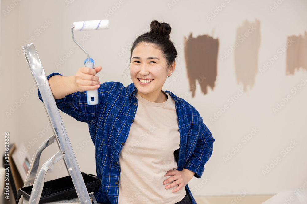A 25-year-old brunette girl with gathered hair, brown eyes and a cute smile holds a paint roller in her right hand. She is leaning on a ladder that she uses during repairs. A 25-year-old brunette girl with gathered hair, brown eyes and a cute smile holds a paint roller in her right hand. She is leaning on a ladder that she uses during repairs.
