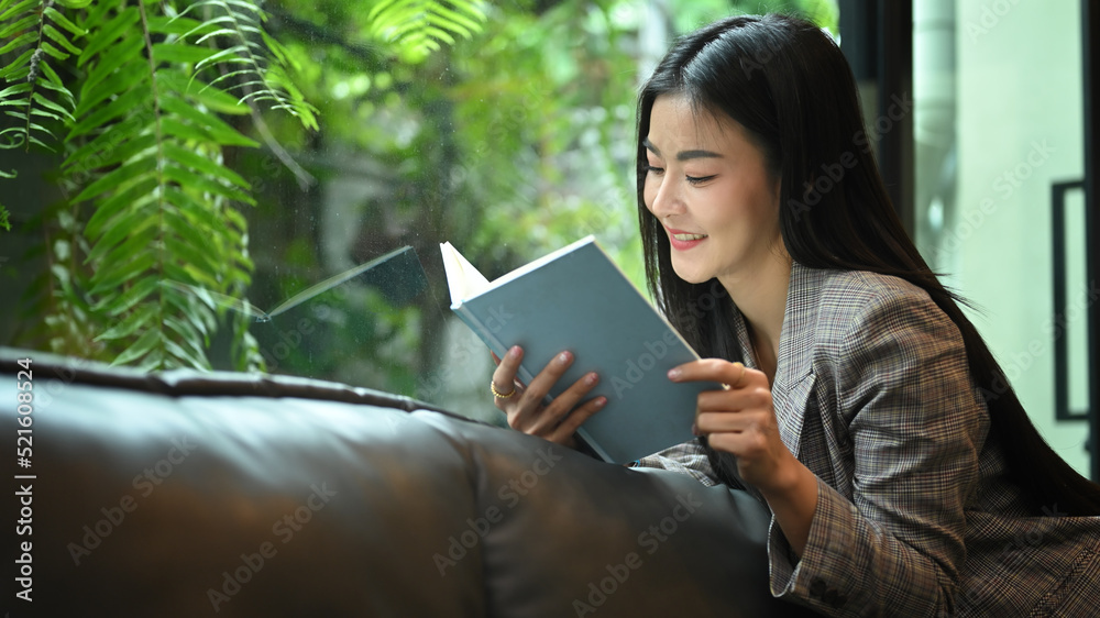 Fototapeta premium Relaxed working woman resting, taking a break from work and reading a book on sofa