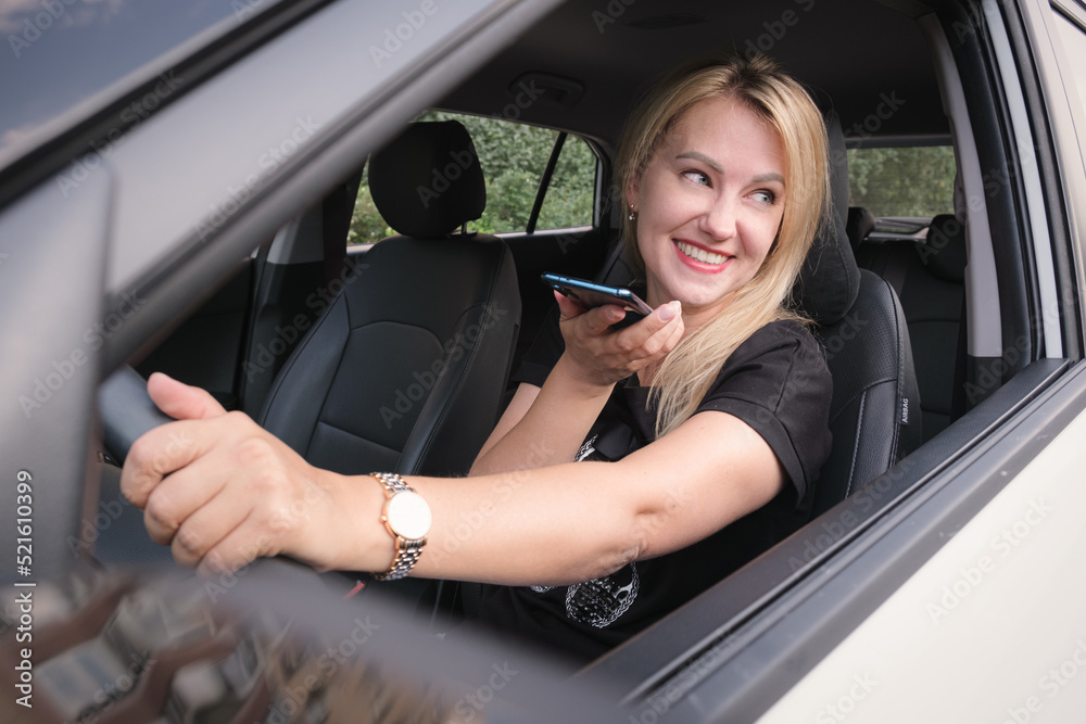 Beautiful woman driving a car is talking on mobile phone and smiling. Woman is holding smartphone and talking on the speakerphone. Blonde holds on to the steering wheel of car and looks to the side