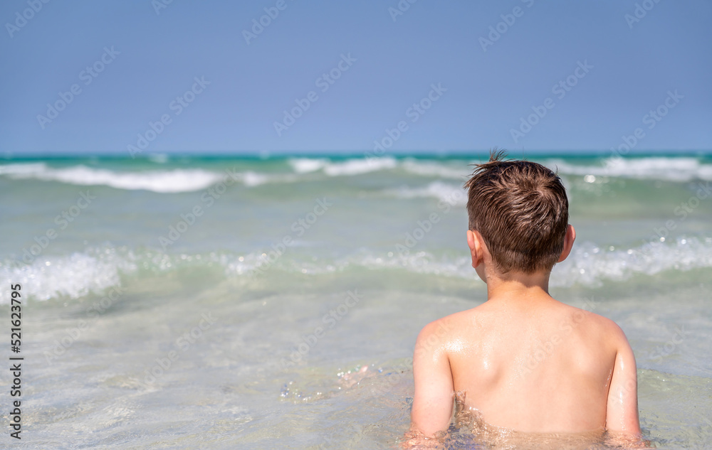 Junge badet in der fachen Brandung eines türkisblauen Meeres vor blauem Himmel Junge badet in der fachen Brandung eines türkisblauen Meeres vor blauem Himmel
