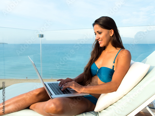 Beautiful young woman sunbathing beside swimming pool and using notebook