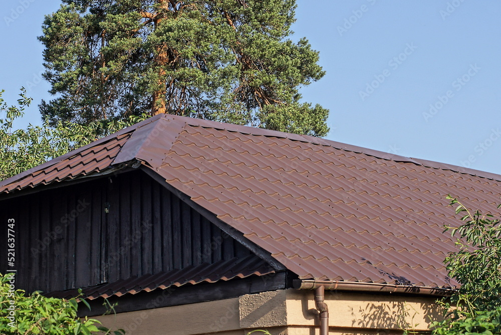 attic of a wooden rural house under a brown tiled roof on the street against the background of a blue sky and a green pine tree attic of a wooden rural house under a brown tiled roof on the street against the background of a blue sky and a green pine tree