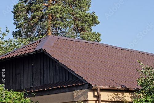 attic of a wooden rural house under a brown tiled roof on the street against the background of a blue sky and a green pine tree © butus