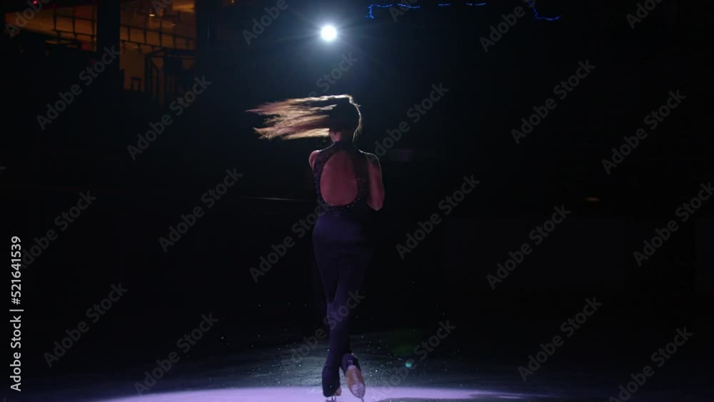 Beautiful young skater girl spin on dark ice rink in spotlight, figure skating performance rehearsal