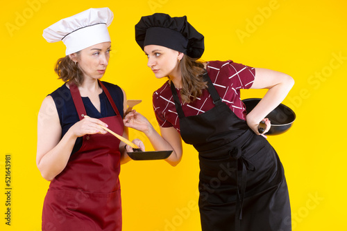Housewives prepare food. Woman in chefs hats. Two female chefs. Woman cook is holding frying pan. Concept sale of kitchen utensils. Housewives cook aprons on yellow background. Cafe chefs girl