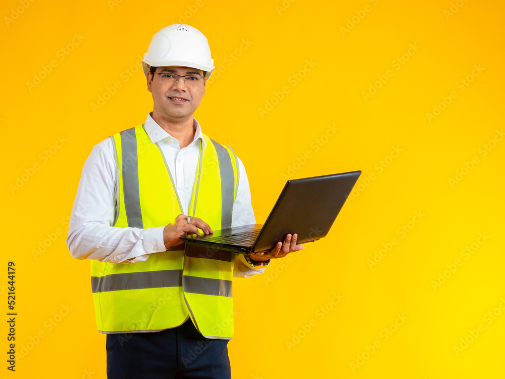 Builder with laptop. Hindu man is standing with computer. Guy in reflective vest on white shirt. Male builder in protective helmet. Architect construction company on yellow background. Builder Career Builder with laptop. Hindu man is standing with computer. Guy in reflective vest on white shirt. Male builder in protective helmet. Architect construction company on yellow background. Builder Career