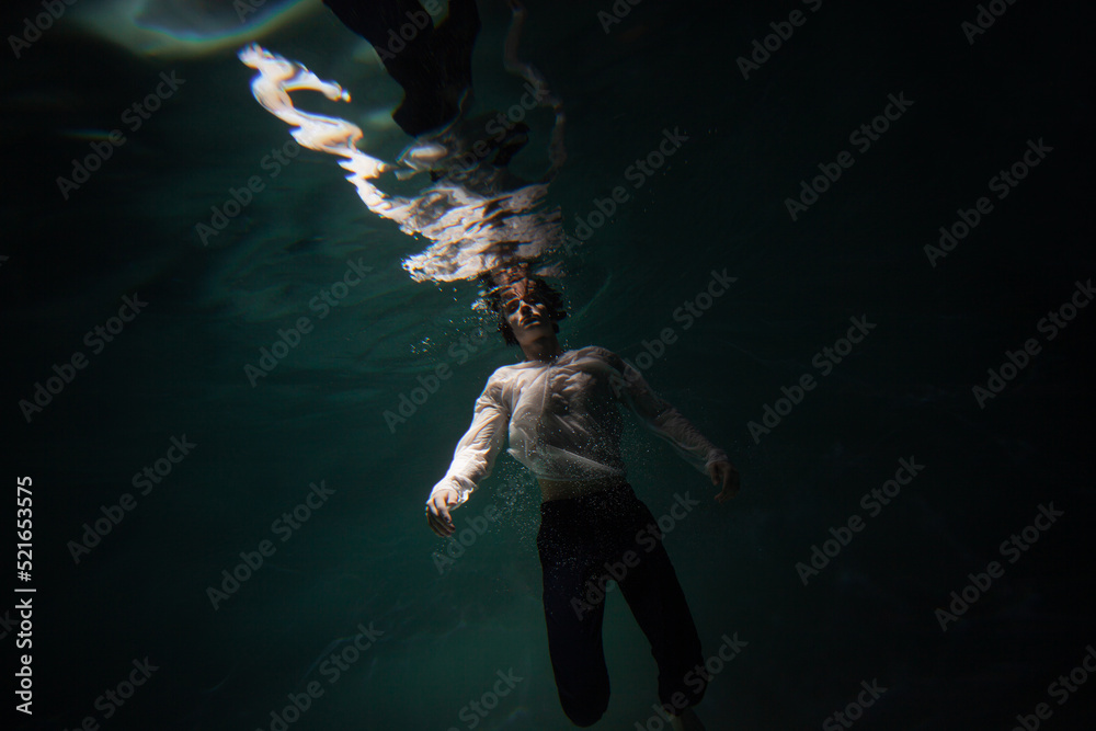 Beautiful underwater shooting, guy in white shirt and pants has fallen under the water and is drowning. a young man relaxes down under surface of the water, waves and refraction of light under water
