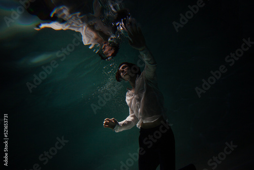 Photo underwater, a guy in a white shirt floundering underwater and reaching for the surface of the water, a man and his reflection. mystical underwater portrait