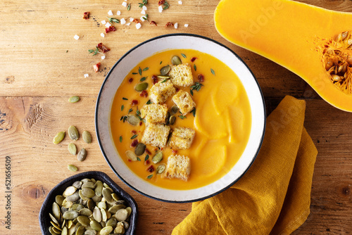 Autumn pumpkin cream soup in bowl served with seeds and crouton on wooden table top view.