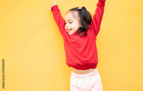 Little girl jumps exited with hands up. She is isolated in a yellow background and wears a red sweater