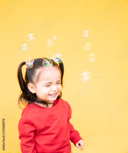 Little girl playing with bubbles. She closes her eyes and smiles tenderly. She is isolated in a yellow background and wears a red sweater