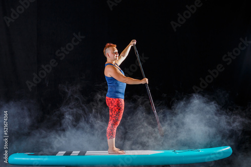 A woman with a mohawk on a sub board with an oar in the fog on a black background.