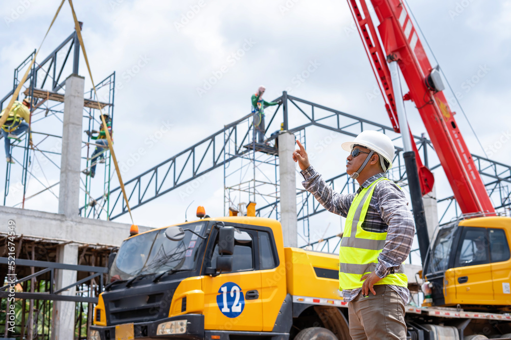 A construction engineer inspects the installation of a factory roof steel frame that uses a mobile crane to lift it up to a height. A construction engineer inspects the installation of a factory roof steel frame that uses a mobile crane to lift it up to a height.